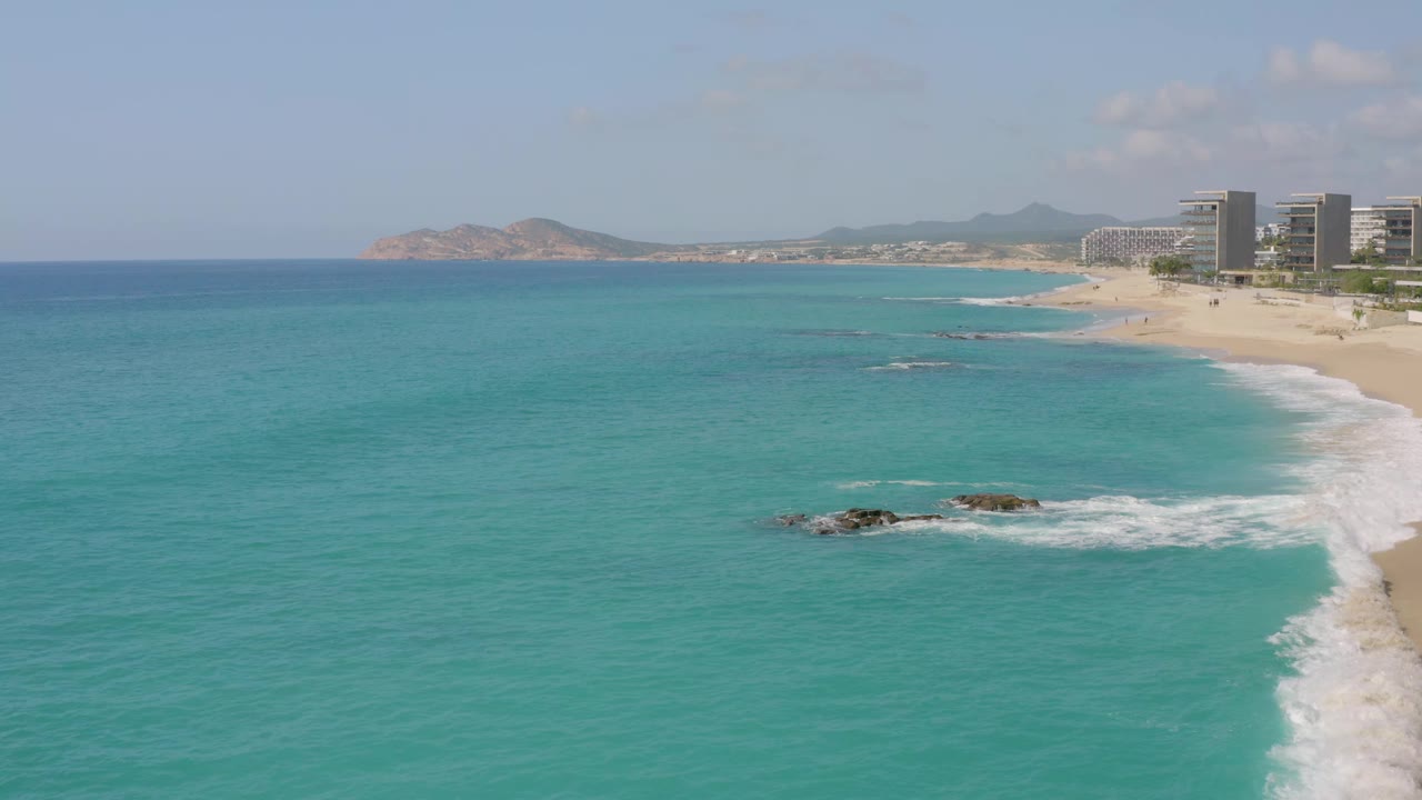 At 0:01, the Dreams Beach shoreline remains in a wide aerial composition, with a dark rock cluster and white water nearer the lower center, beachfront buildings on the right, distant hills, and no visible people.