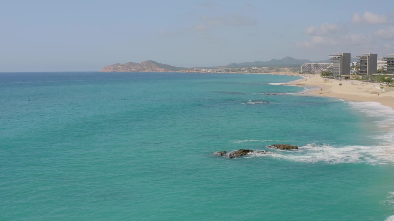 At 0:02, turquoise water dominates the Dreams Beach frame, with several dark rock patches in the midground, a pale beach curving behind them, modern buildings at the far right, hills beyond, and no visible people.
