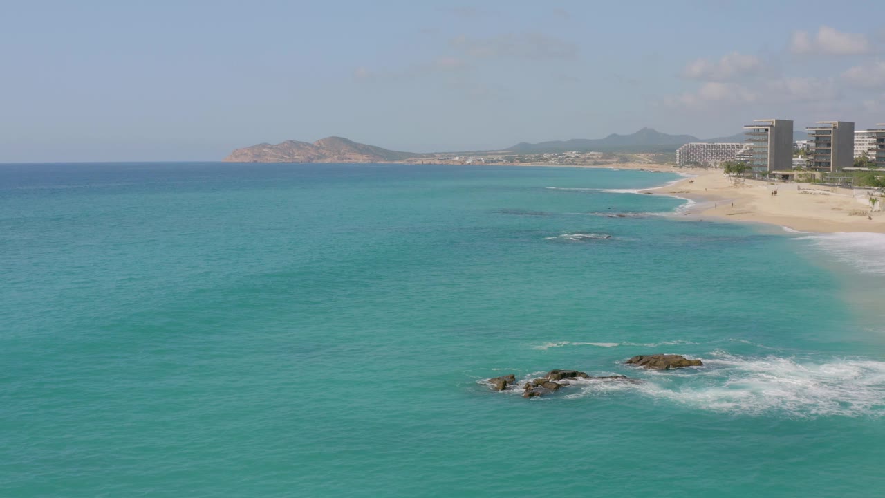 At 0:03, Dreams Beach is seen from above with a larger rock cluster and foamy wave wash low in the frame, open turquoise water to the left, sandy shoreline and buildings to the right, and no visible people.