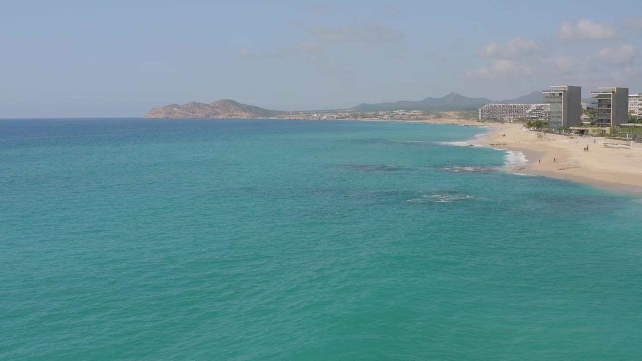 One second into the Dreams Beach aerial, the turquoise water dominates the lower frame while the curving sand and modern resort buildings remain on the right with no visible people.