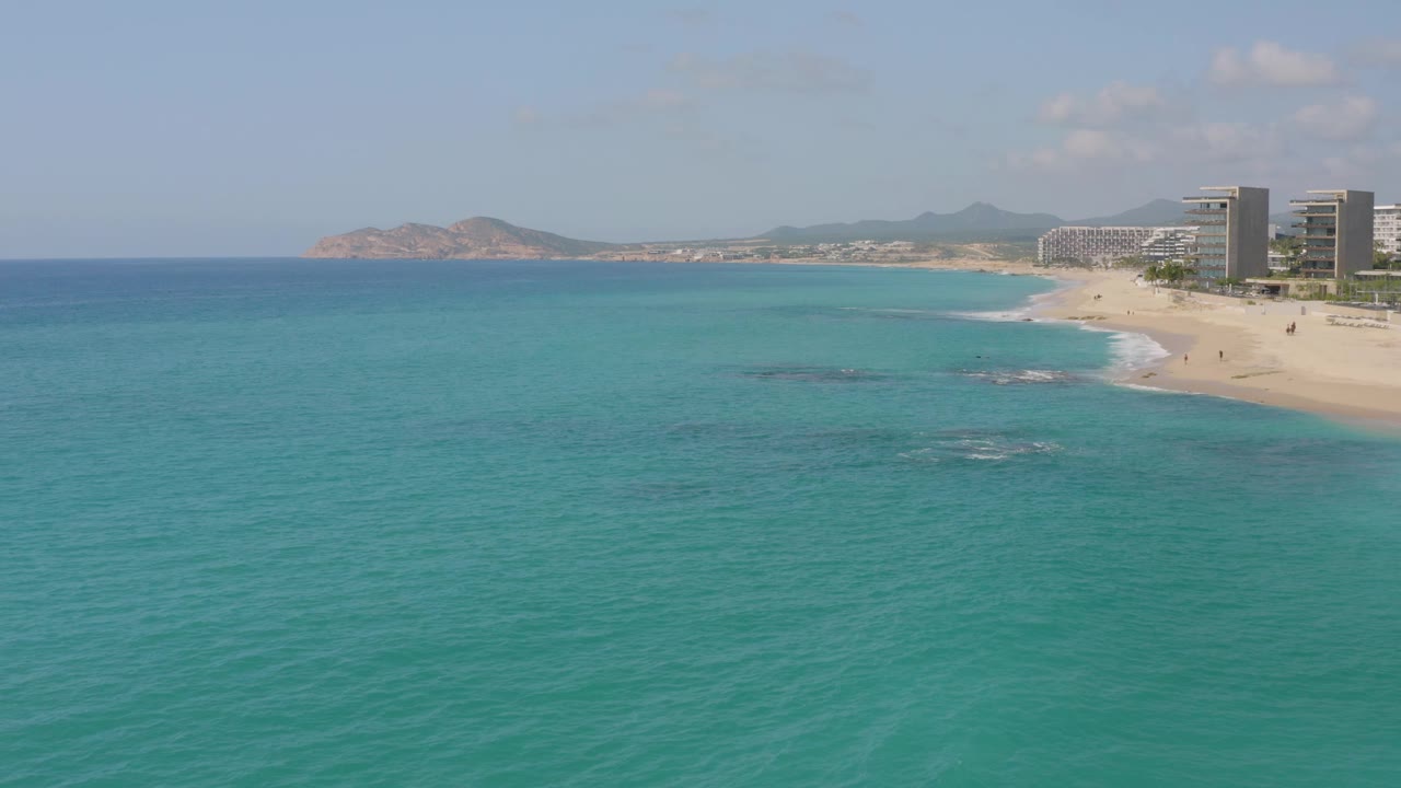 At 0:02, the aerial view shows more open turquoise water in the foreground, shallow darker patches offshore, the Dreams Beach resort buildings at right, and no visible people.