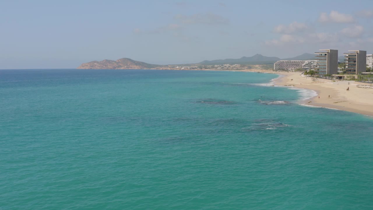 Mid-clip frame over Dreams Beach with the beach running diagonally along the right edge, resort towers beyond it, wide ocean across the foreground, and no visible people.