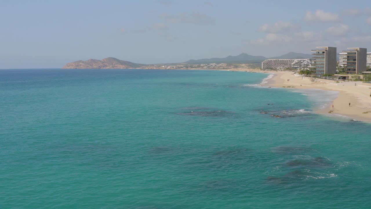 Wide aerial view at Dreams Beach in Baja California Sur, Mexico, with turquoise water in front, pale sand and hotels at right, distant hills on the horizon, and no visible people.
