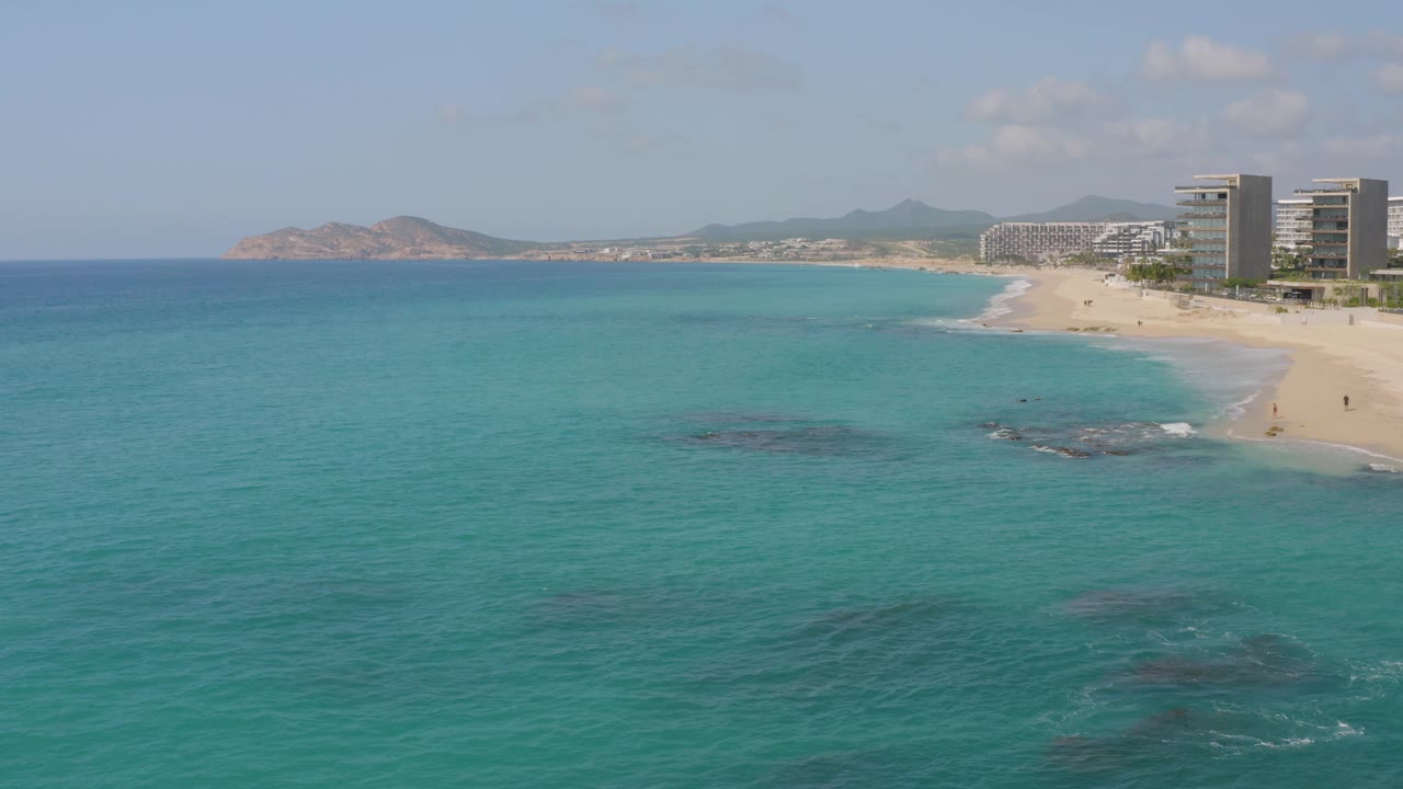 High coastal view at Dreams Beach in Baja California Sur, Mexico, with a broad turquoise ocean foreground, a curving sandy beach, hotel towers to the right, distant hills, and no visible people.