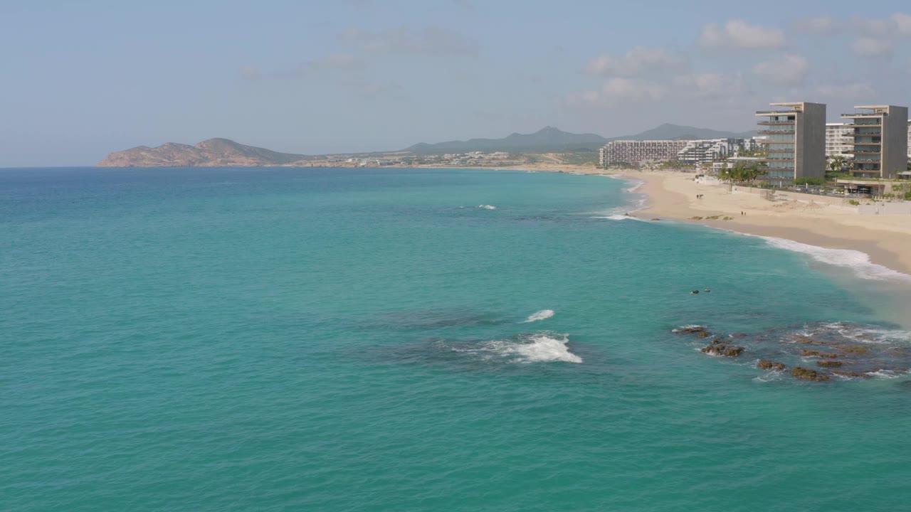 Wide aerial frame at Dreams Beach, Baja California Sur, Mexico, showing blue-green water across the foreground, small white wave breaks over rocks, resort towers along the right beach edge, distant hills, and no visible people.