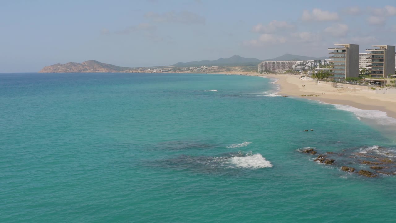 Drone-height view of Dreams Beach in Baja California Sur, Mexico, with turquoise water across the frame, a larger white surf break over dark rocks near the lower right, resort buildings on the beach, distant coastline, and no visible people.