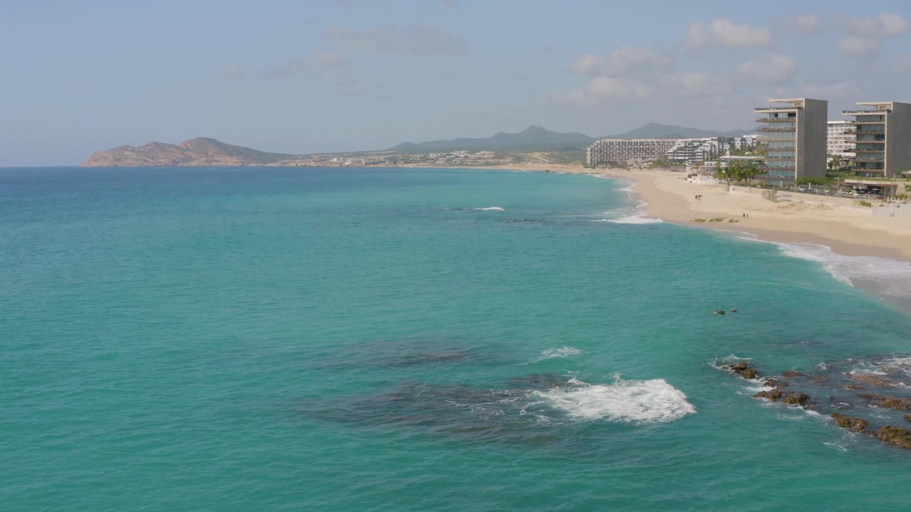 High coastal view with open turquoise sea, sandy shore, resort buildings, and rocky surf.