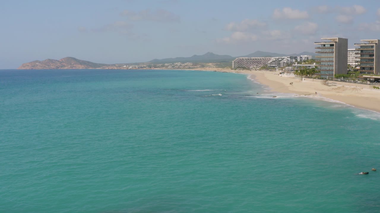 At Dreams Beach in Baja California Sur, Mexico, turquoise water fills the foreground while a sandy beach, beachfront towers, distant hills, and no visible people sit along the right side.