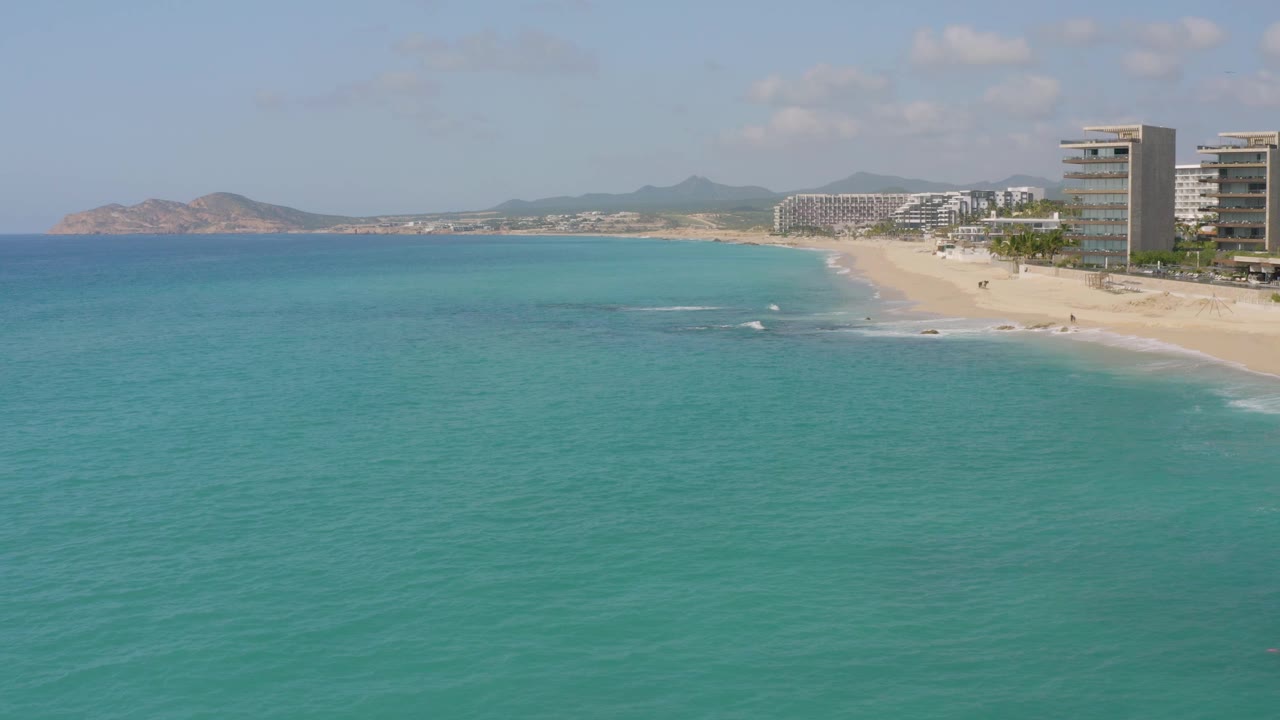 At Dreams Beach in Baja California Sur, Mexico, the frame shows a broad blue sea, a curving sandy beach, hotel-like buildings near the shore, hazy hills beyond, and no visible people.