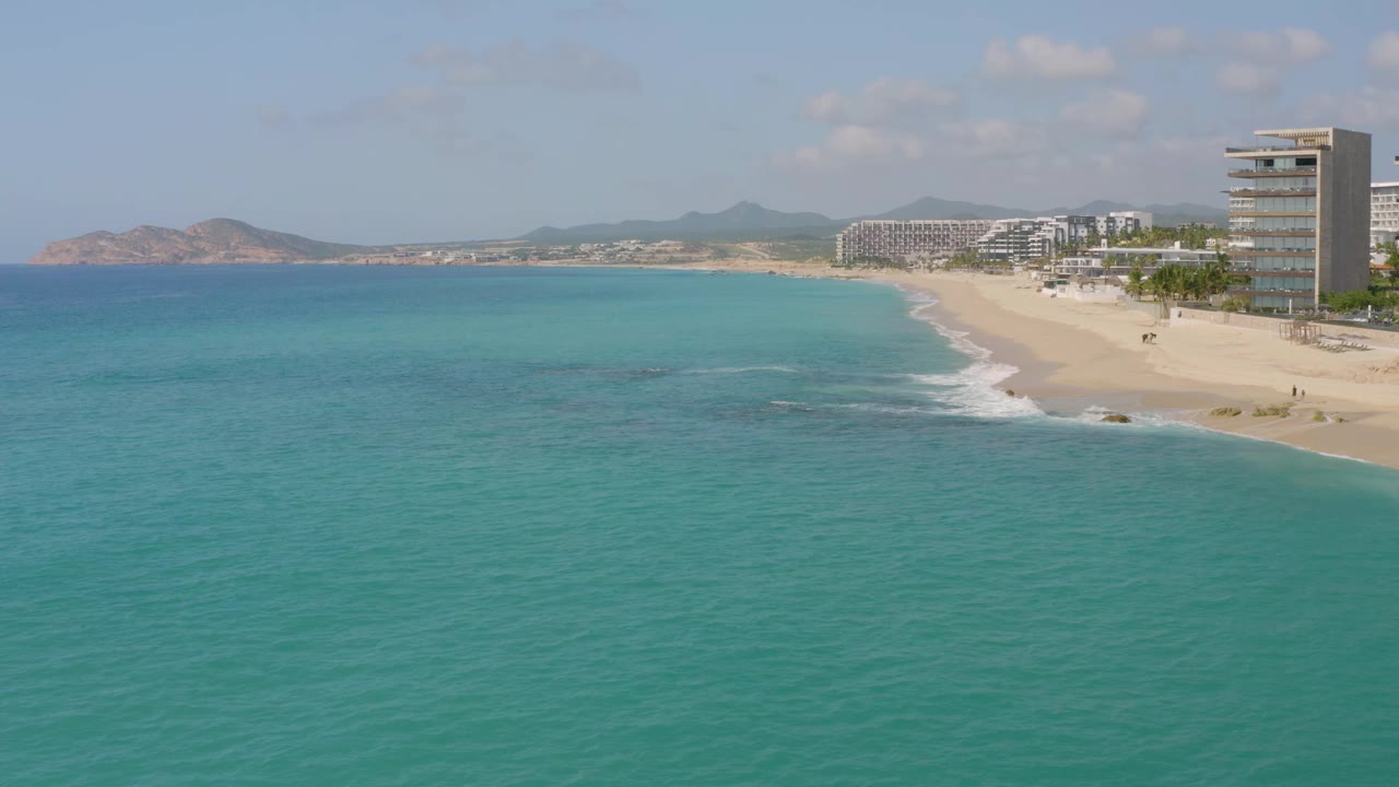 Aerial view one second into the clip shows Dreams Beach with blue-green water covering most of the frame, a pale sand shoreline on the right, resort buildings in the background, low mountains, and no visible people.
