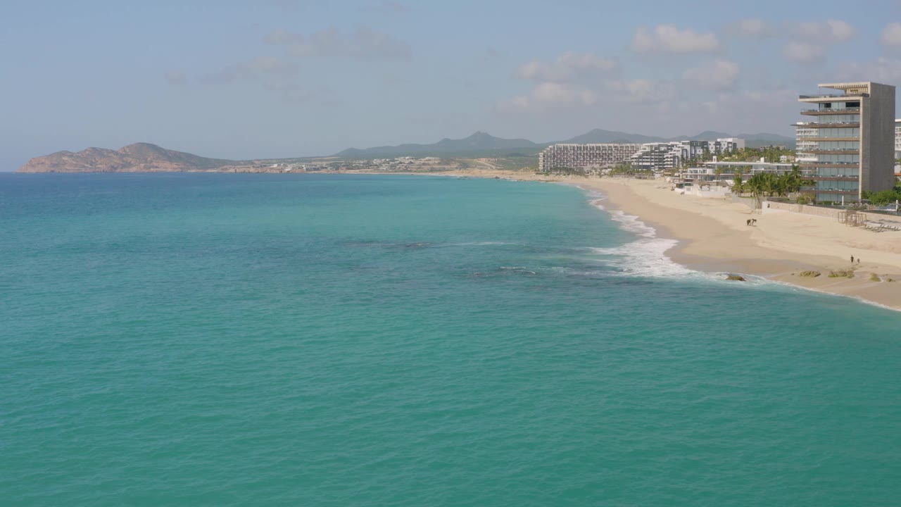 Frame two shows the Dreams Beach shoreline from above, with turquoise sea in front, a curving surf line, sandy beach on the right, modern beachfront buildings behind it, distant terrain, and no visible people.