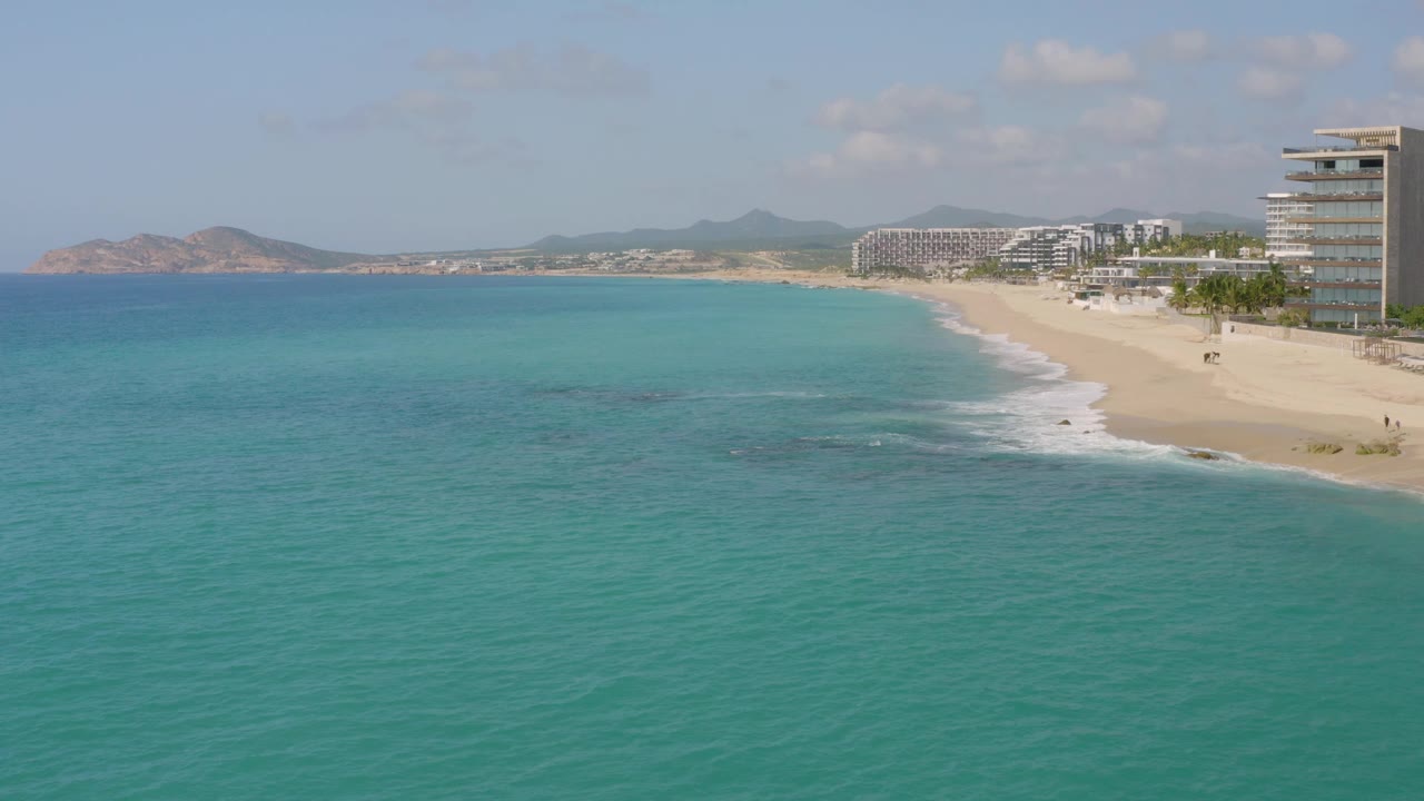 At three seconds, the aerial composition places the ocean in the foreground, the white shore break along the sand, beachfront buildings to the right, hills across the horizon, and no visible people.