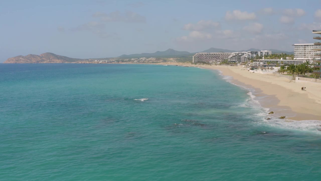 Aerial frame at Dreams Beach, Mexico, showing broad blue water, a curving sandy shoreline, white surf lines, hotel-style buildings to the right, distant hills, and no visible people.