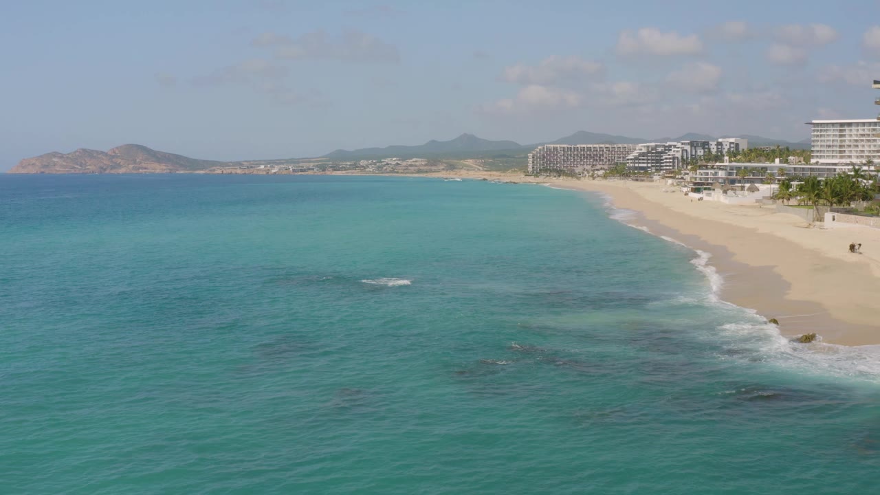 Drone view over Dreams Beach in Baja California Sur with clear blue-green water filling most of the frame, beach and resort buildings along the right edge, hills on the horizon, and no visible people.