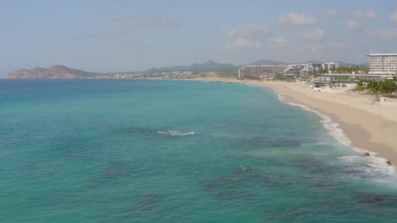 Mid-clip aerial frame at Dreams Beach, Mexico, with turquoise water and a small white wave patch in front, sandy beach and buildings to the right, low hills beyond, and no visible people.