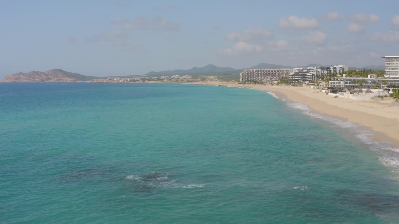 At Dreams Beach in Baja California Sur, turquoise water fills the foreground while a sandy beach, resort buildings, and distant low hills sit under bright sky, with no visible people.