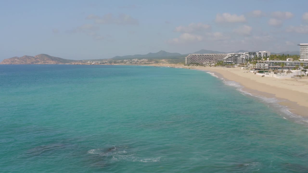 A wide offshore view of Dreams Beach shows turquoise water spreading across most of the frame, a curving sand line to the right, resort buildings behind it, and no visible people.