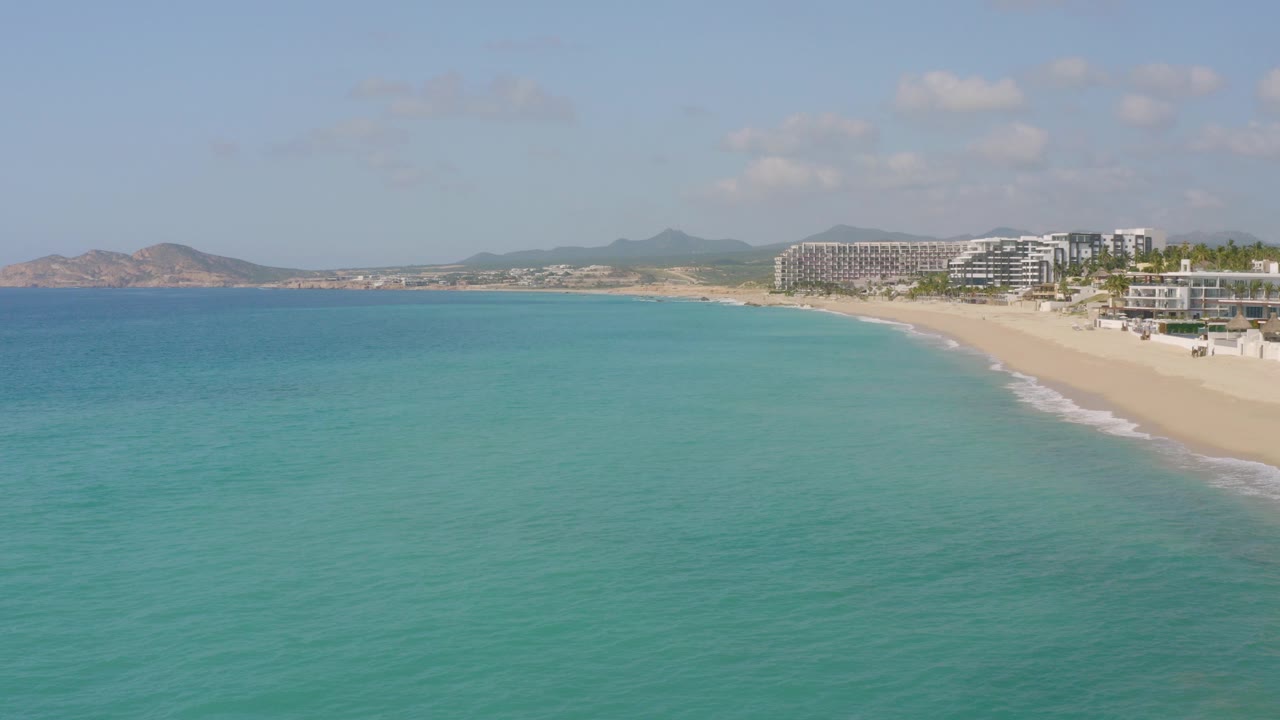 Opening frame of Dreams Beach in Baja California Sur, Mexico, showing turquoise water in the foreground, a long sandy shoreline on the right, distant hills, beachfront buildings, blue sky, and no visible people.