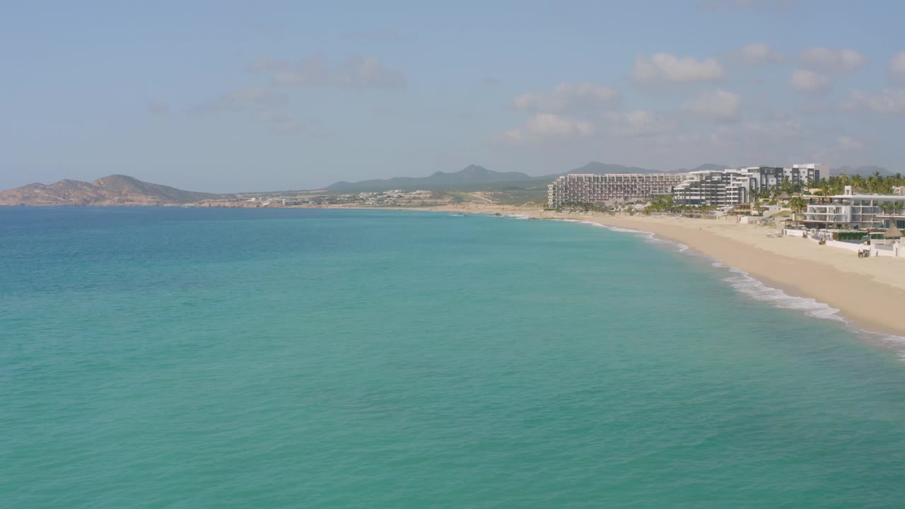 At 0:02, the aerial view over Dreams Beach shows blue-green water, a gently curving sand strip, a thin white surf edge, modern beachfront structures, hazy hills, and no visible people.