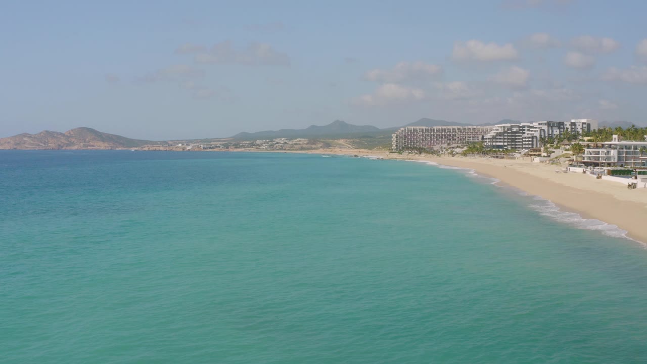 At 0:03, Dreams Beach remains in a wide coastal composition with turquoise water across the foreground, pale sand along the right, distant landforms to the left, beachfront buildings, and no visible people.