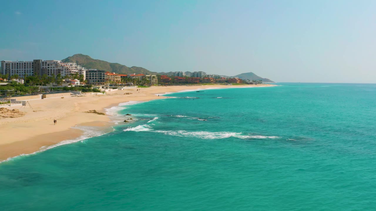 Aerial view of Dreams Beach with a small line of surf reaching the sand, clear blue-green water across the foreground, hotel buildings at the left, distant hills, and no visible people.