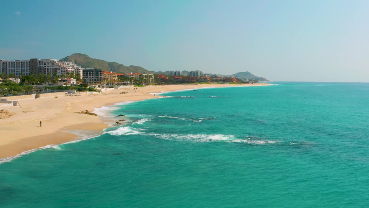 The frame holds a broad turquoise sea beside Dreams Beach, where thin white waves cross shallow rock patches near the sand and beachfront buildings sit below dry hills, with no visible people.