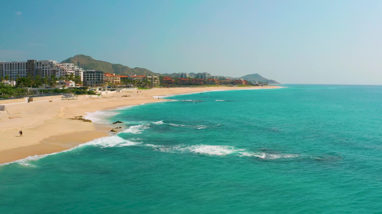 Mid-clip aerial frame over Dreams Beach showing foam around nearshore rocks, a wide empty-looking sand strip, resort-style buildings along the back of the beach, hills behind them, and no visible people.