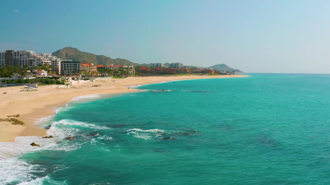 Aerial frame at Dreams Beach showing white surf curling onto the sand, dark rocks just offshore, turquoise water across the right side, coastal buildings at left, and no visible people.
