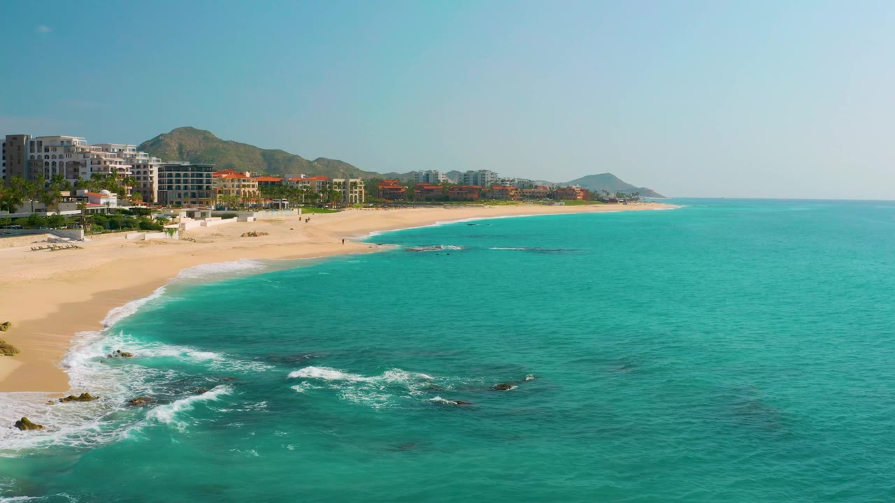 Aerial view of Dreams Beach with more open turquoise water filling the right side, pale sand sweeping from the left foreground into the distance, resort-style buildings and hills behind it, and no visible people.