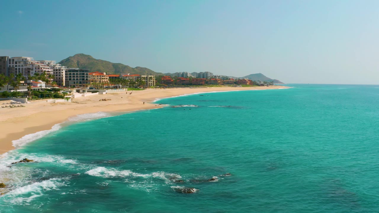 Mid-clip aerial frame at Dreams Beach with foamy surf in the lower left, scattered dark rocks in the nearshore water, a long sandy shoreline, background buildings, hills, and no visible people.