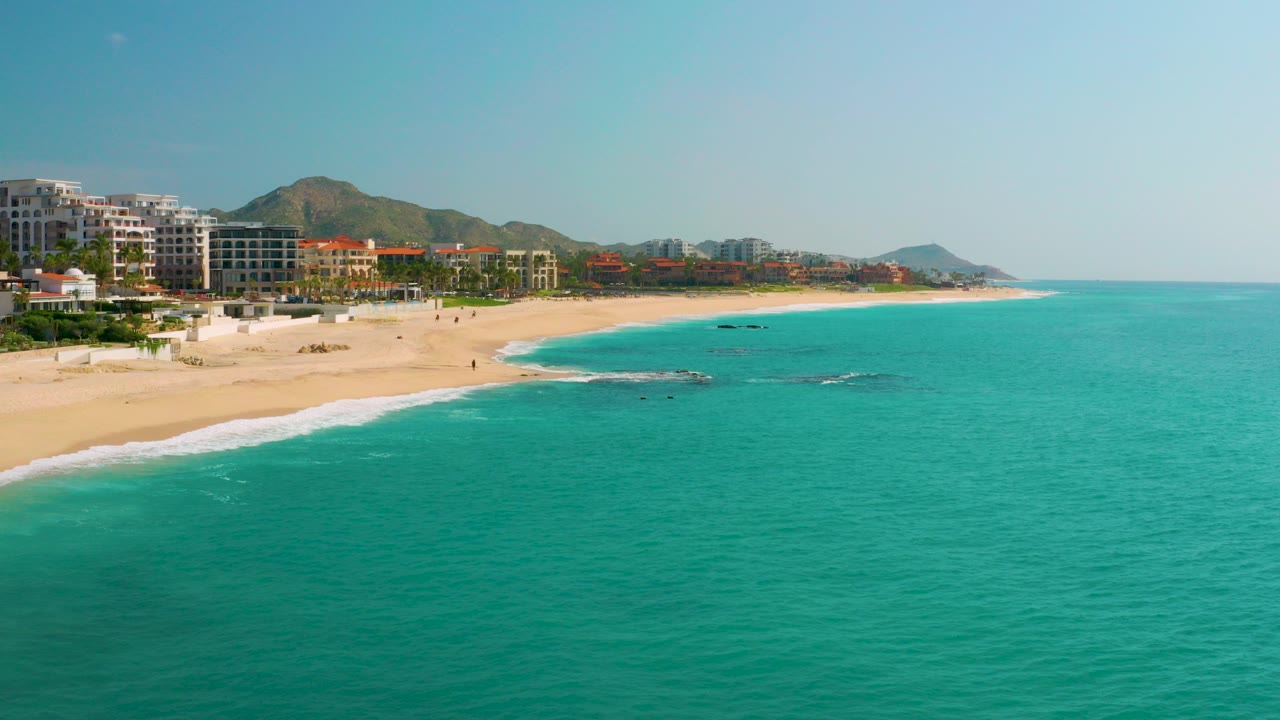 Aerial view of turquoise ocean beside a sandy beach with beachfront buildings and hills.