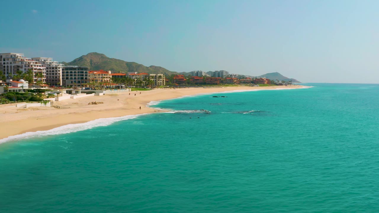 Aerial view at Dreams Beach, Baja California Sur, Mexico, with the shoreline curving from left to center, small surf lines, offshore rocks, hillside background, and no visible people.