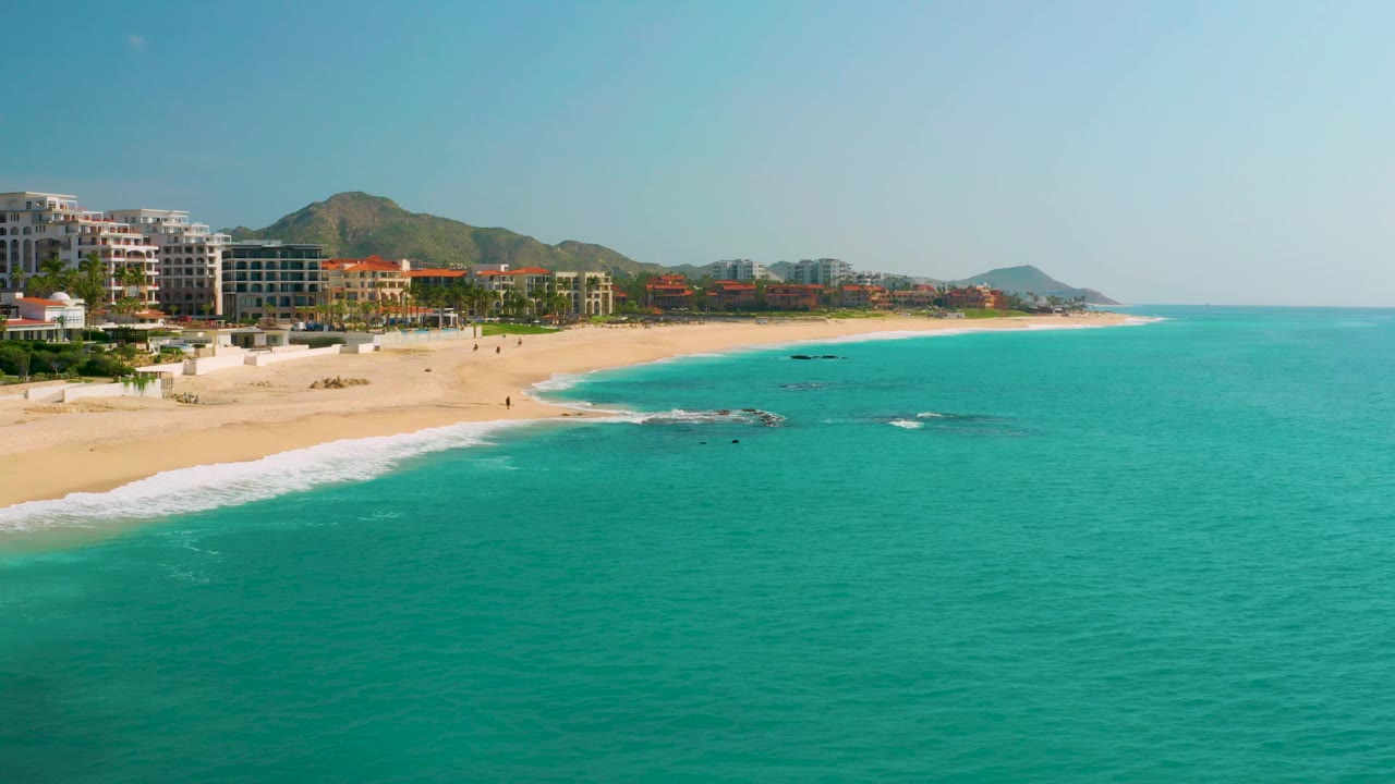 Elevated frame from Dreams Beach in Baja California Sur, Mexico, showing more open turquoise sea to the right, pale sand on the left, low surf near rocks, and no visible people.