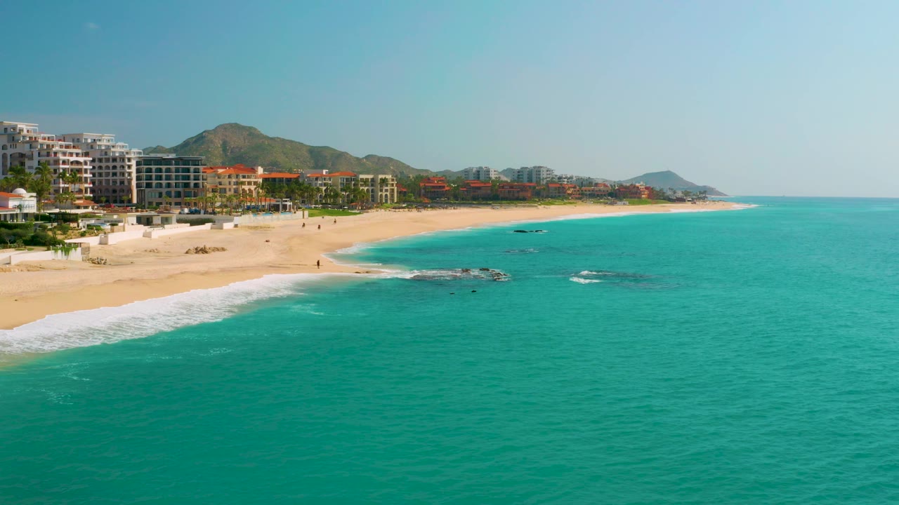 Aerial frame over Dreams Beach, Baja California Sur, Mexico, with beachfront buildings tighter on the left, a pale beach through the center, turquoise water across the foreground, and no visible people.