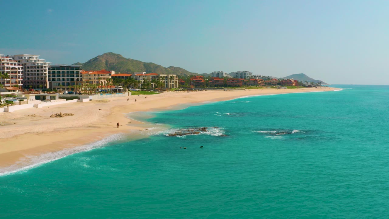 Opening aerial frame over Dreams Beach in Baja California Sur, Mexico, with turquoise water, a curving sand shoreline, hotel buildings, rocky surf, distant hills, and no visible people.