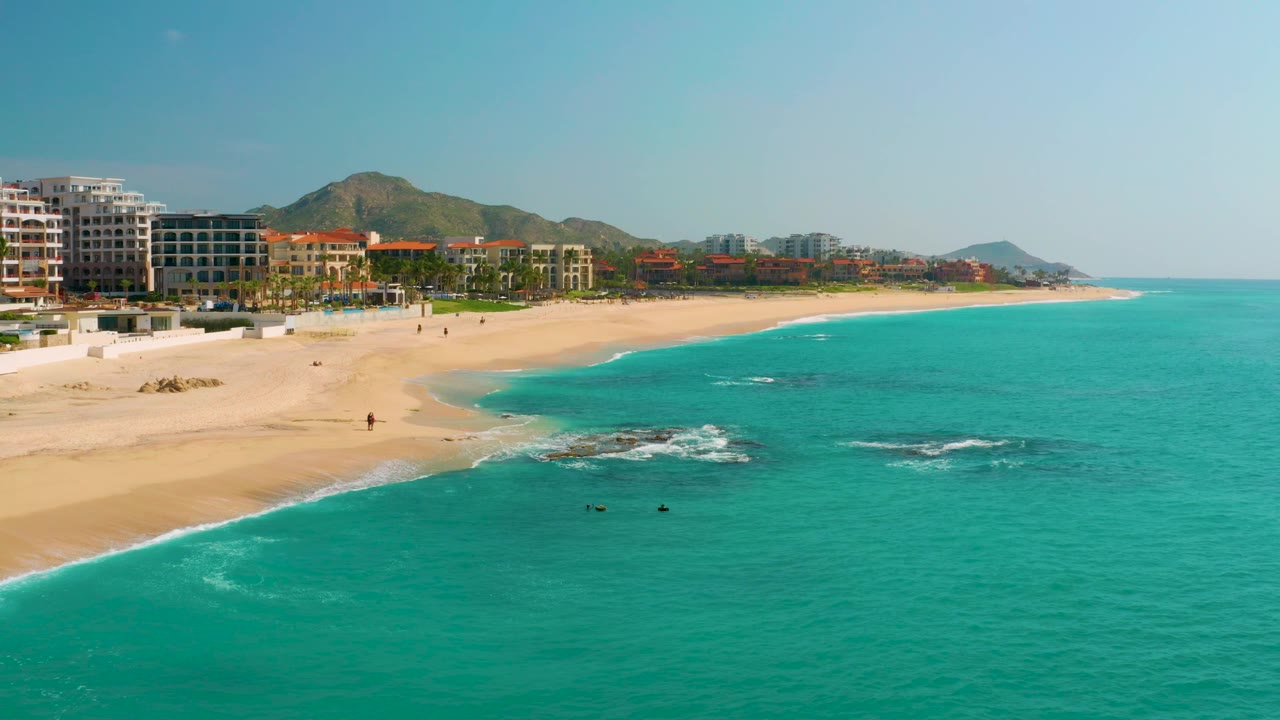 High coastal view at Dreams Beach in Baja California Sur, Mexico, with more open water to the right, a sandy beach sweeping left to center, small surf over rocks, hotel blocks, distant hills, and no visible people.