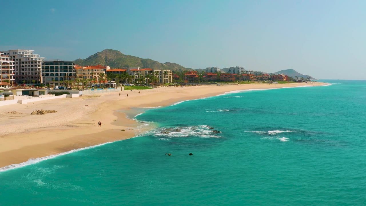 Aerial coastline view with beach, hotel buildings, rocks in the surf, and turquoise sea.