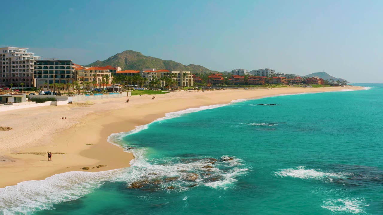 Elevated shoreline frame at Dreams Beach in Baja California Sur, Mexico, showing surf wrapping around dark rocks near the lower center, a broad sandy beach to the left, turquoise water to the right, resort buildings, palms, hills, and no visible people identifiable.