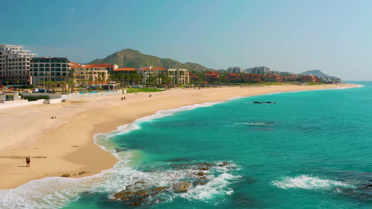 A Dreams Beach aerial in Baja California Sur, Mexico, with the shoreline curve more centered, white foam crossing the lower beach edge, rocks breaking the water near the bottom, resort buildings and palms behind the sand, distant hills, and no visible people identifiable.