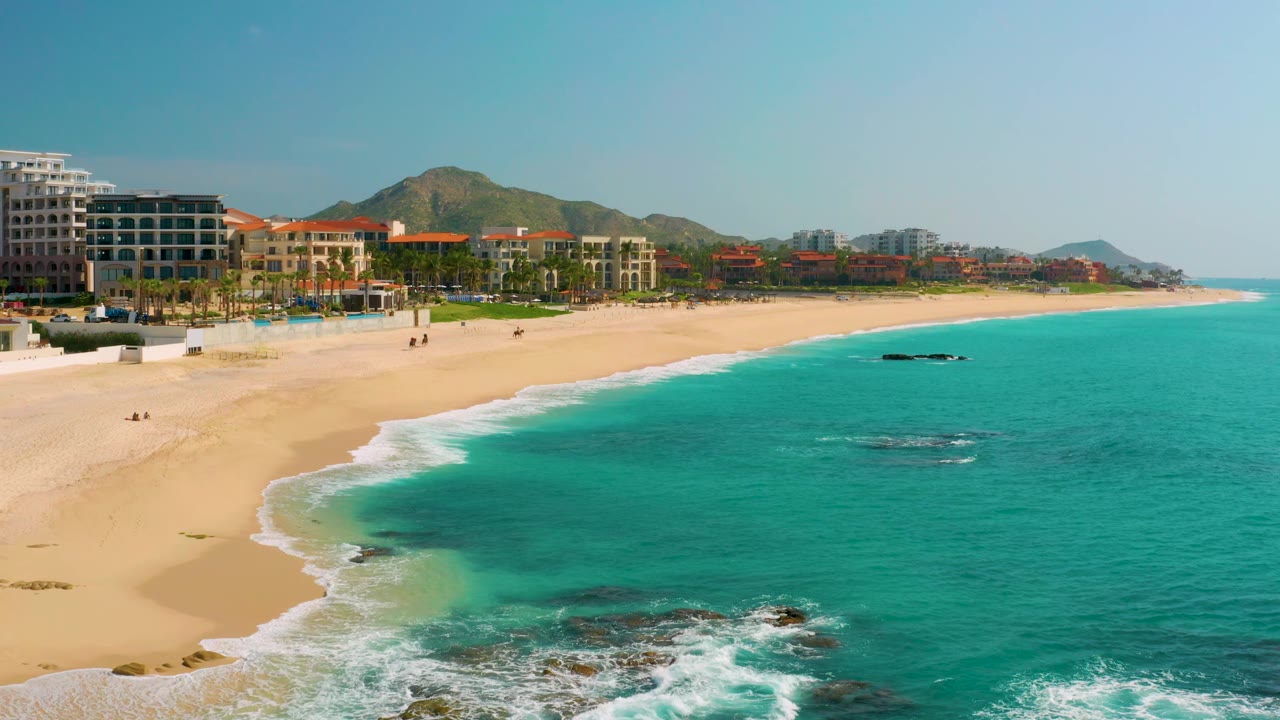 Beach resort coastline with turquoise water, sandy shore, small rocks, and distant hills.