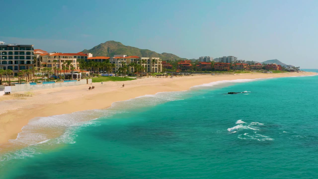 Wide beach curve with resort buildings, palms, blue water, and distant mountains.
