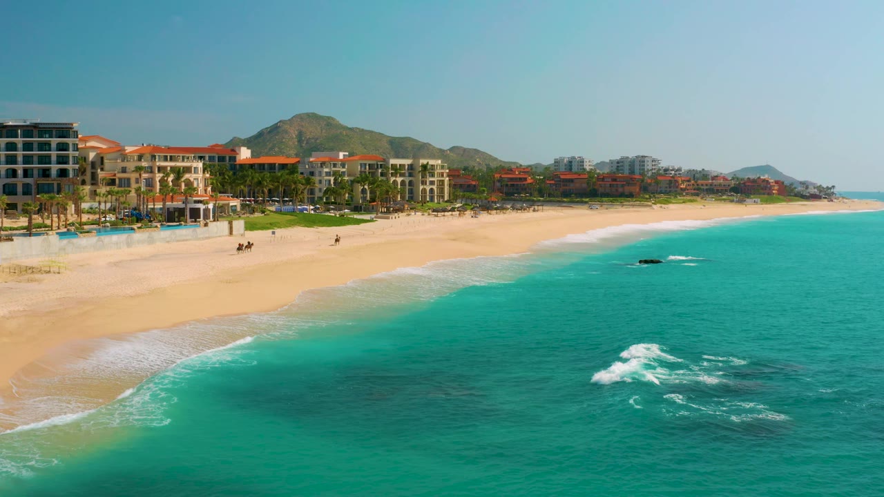 Aerial frame at Dreams Beach showing white surf over darker shallow rocks, a broad sand strip, resort buildings, palm trees, and mountains in the background.