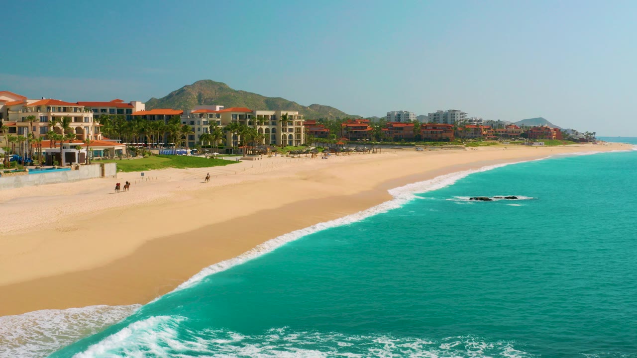 Wide aerial frame from Dreams Beach in Baja California Sur showing turquoise sea in the foreground, a pale sandy shoreline, beachfront buildings with red roofs, low mountains beyond, and no visible people.