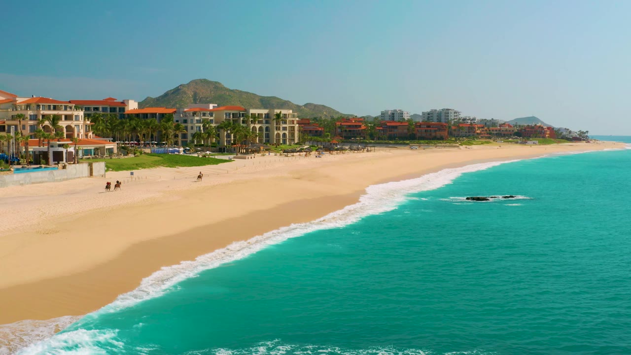 Dreams Beach appears from above with the waterline pushed slightly higher on the sand, a small dark rock in the turquoise water, beachfront buildings and palms to the left, and no visible people.