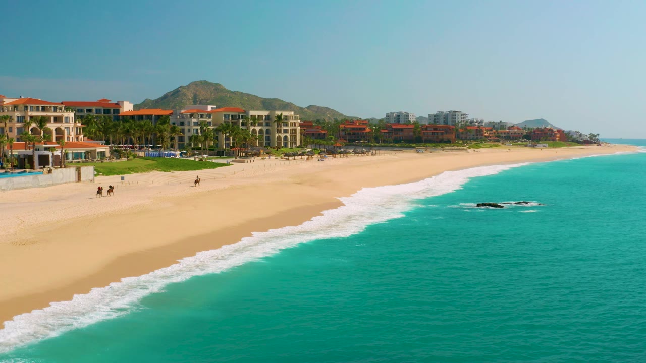 At 0:03, the aerial view over Dreams Beach shows a longer stretch of white wash along the sand, turquoise water across the lower right, beachfront buildings at upper left, mountains behind, and no visible people.