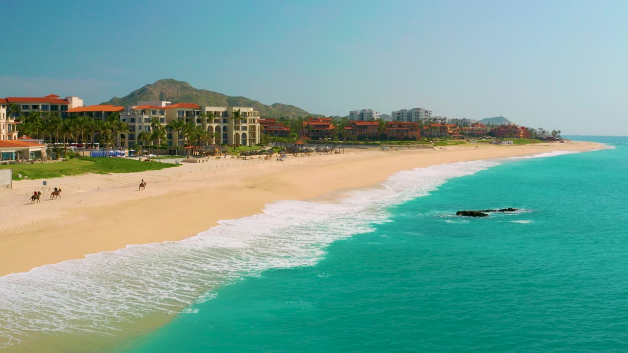 Wide aerial frame at Dreams Beach in Baja California Sur, Mexico, with turquoise surf in the foreground, a broad sand beach, beachfront buildings, palms, and low hills beyond.