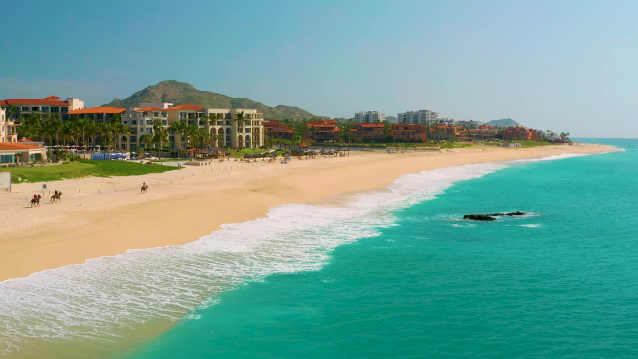 At 0:01, the Dreams Beach shoreline remains wide in frame, with foamy waves reaching the sand, beachfront buildings to the left, and tiny distant figures on the beach.