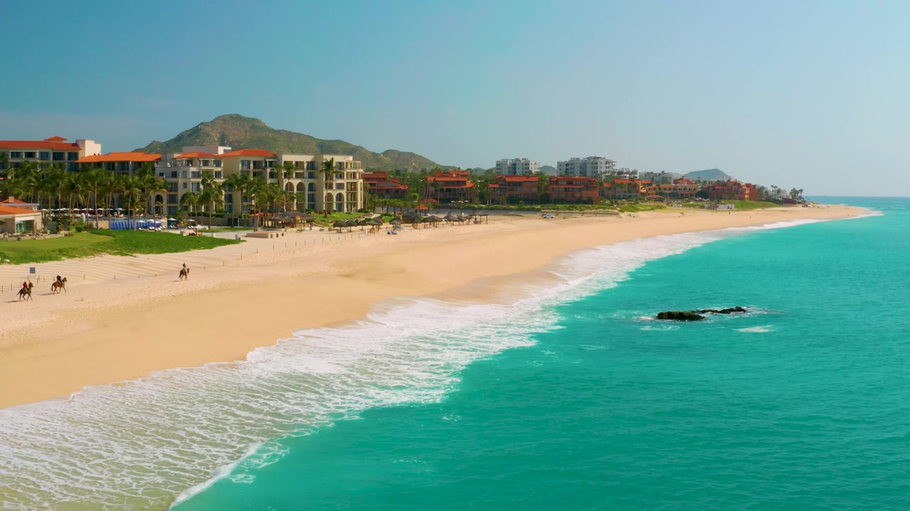 At 0:02, the camera view shows a longer stretch of Dreams Beach, with turquoise water at right, a white surf edge, dark rocks offshore, and buildings running along the sand.
