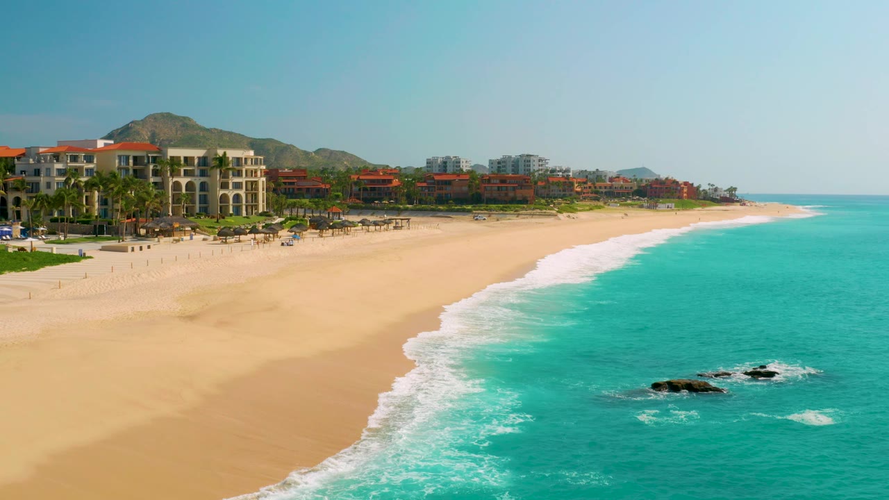 At 0:02, the coastline stretches farther right with resort buildings and palms on the left, open sand in the center, blue-green water on the right, and no visible people.