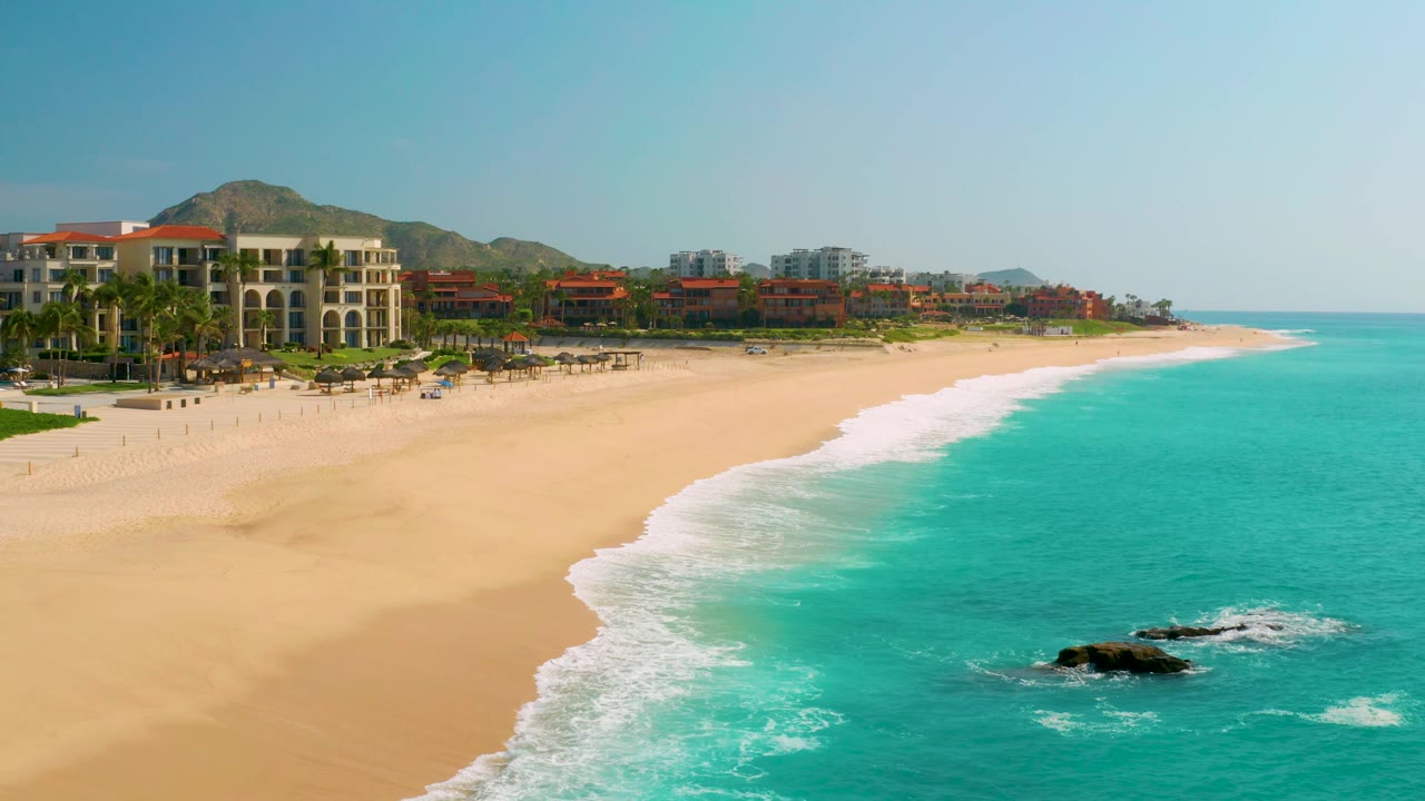 Elevated view of resort-lined beach with surf wrapping around dark offshore rocks.